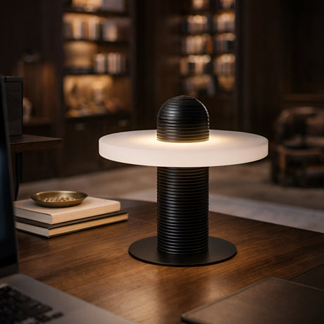 The Nyxara LED Single Tiered Alabaster Disc Table Lamp in soft white and black sits on a wooden desk in a cozy, book-filled study, glowing warmly against shelves of books and ambient lighting.
