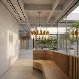 A modern, minimalist interior with a curved wooden bench, large windows, and the Tycho 6 Light Crystal Chandelier in gold and white, offering views of a green courtyard filled with trees and natural light.