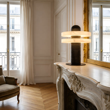 Elegant living room with tall windows, sheer curtains, and an ornate white marble fireplace. The Nyxara LED Double Tiered Alabaster Table Lamp in Soft White & Black adds a modern touch, while a gold-framed mirror and armchair complete the sophisticated space.