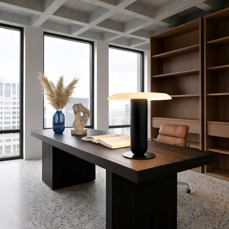 Modern office with large windows, a dark wood desk displaying the Luxelle LED Single Tiered Alabaster Table Lamp in soft white & brass, an open book, abstract sculpture, and blue vase with pampas grass; shelves and a brown leather chair in the background.