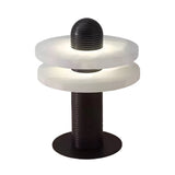 The Nyxara LED Double Tiered Alabaster Table Lamp features a cylindrical black base, two frosted white rings separated by a gap, and a grooved black column. Lit in soft white and black, it creates a modern, glowing ambiance.