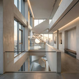 A modern, minimalist interior with high ceilings, matte white walls, and a sculptural spiral staircase features the Universe LED Suspension Light - Matte White. Large windows reveal sunset-lit city buildings while contemporary fixtures add elegance.