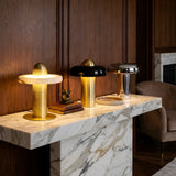 Three stylish modern table lamps—including the Nyxara LED Single Tiered Alabaster Table Lamp in Soft White & Brass—shine on a marble console against a wood-paneled wall, next to stacked books and a small sculpture.