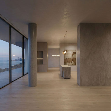 Minimalist modern interior with light wood floors, concrete walls, and a central pillar. Floor-to-ceiling windows offer ocean and city views. The Luna LED Linear Suspension Light in gold elegantly illuminates the sleek island for a refined touch.