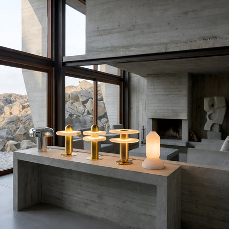 A modern concrete interior with large windows, featuring the Nyxara LED Double Tiered Alabaster Table Lamp in Soft White & Brass and other decorative lamps on a minimalist console table. Rocks are outside and abstract art decorates the background.