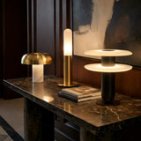 Three modern table lamps, including the Luxelle LED Double Tiered Alabaster Table Lamp in Soft White & Black, are displayed on a dark marble console with stacked books, set against a wood-paneled wall and abstract artwork in a chic interior.