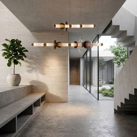 A modern minimalist interior with concrete floors and walls features a potted plant on a bench, geometric pendant lights, a Luna LED Linear Double Suspension Light - Gold above, a glass wall to greenery, and an open staircase on the right.