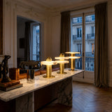 A marble table with the Nyxara LED Single Tiered Alabaster Table Lamp in Soft White & Brass and books sits by a window in an elegant room with parquet floors, intricate wall detailing, and glowing cityscape views.