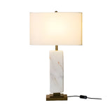 The Brio 1 Light Solid Marble Table Lamp in white and brass features a rectangular marble base, gold accents, a large white fabric shade, and a visible black power cord with switch. The lamp is on, casting warm light.
