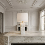 The Kavryn 1 Light Raw Cut Alabaster Table Lamp in brass and soft white with a rectangular shade sits on an ornate marble desk in an elegant room featuring paneled walls, intricate ceilings, large windows, and a fireplace.