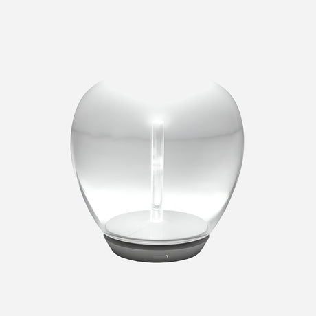 The Empatia LED Table Lamp - Clear is a contemporary, transparent hand-blown glass lamp with a sleek oval shape seated on a simple round base. It emits a soft, white light from its center, presenting a minimalist and elegant look against a plain gray backdrop.