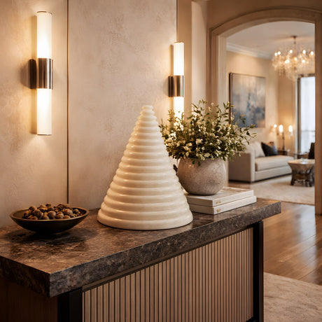 A decorative console table displays a modern white cone sculpture, a round bowl of nuts, a potted plant on books, and the Virelle LED Pyramid Alabaster Table Lamp in Soft White brightening a cozy hallway to the living room.
