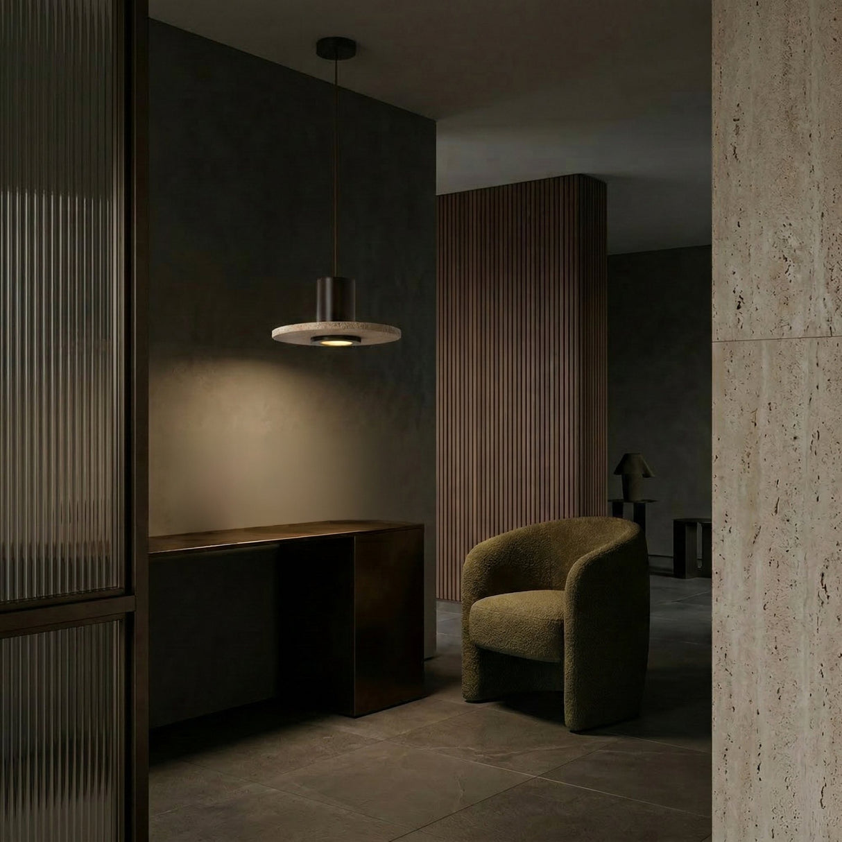 A modern, dimly lit interior showcases a curved olive-green armchair, a dark minimalist desk, and the Torvane 1 Light Small Wide Disc Travertine Pendant Light in Walnut Brown & Light Beige above textured stone walls with vertical wood paneling for a cozy, sophisticated vibe.