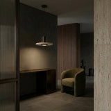 A modern, dimly lit interior showcases a curved olive-green armchair, a dark minimalist desk, and the Torvane 1 Light Small Wide Disc Travertine Pendant Light in Walnut Brown & Light Beige above textured stone walls with vertical wood paneling for a cozy, sophisticated vibe.
