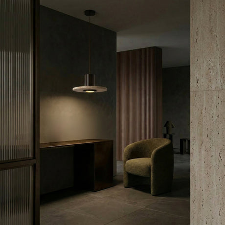 A modern, dimly lit interior showcases a curved olive-green armchair, a dark minimalist desk, and the Torvane 1 Light Small Wide Disc Travertine Pendant Light in Walnut Brown & Light Beige above textured stone walls with vertical wood paneling for a cozy, sophisticated vibe.