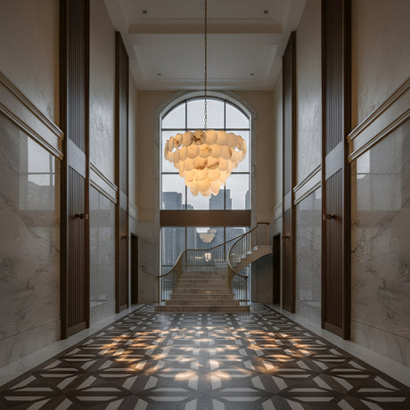 A grand hallway with marble walls, a geometric patterned floor, and the Solyra 21 Light 5 Tier Petal Alabaster Chandelier in soft white. A sweeping staircase curves up to a tall window framing the city skyline.