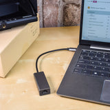 AV:Link USB-C to RJ45 Gigabit Ethernet adaptor by AVSL in brushed aluminium, plugged into a laptop's USB-C port on a wooden desk.