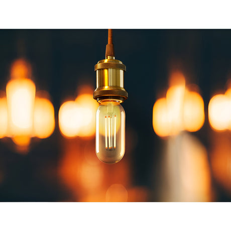 Schuller Vintage T45 8W E27 LED filament bulb in clear glass, suspended from a brass lampholder against a backdrop of warm glowing bokeh lights.