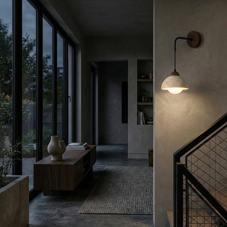 A modern, dim hallway with concrete walls features the Marvoro 1 Light Hanging Bowl Travertine Wall Light in walnut brown and light beige, a wooden bench with vases, large windows facing a garden, carpeted floor, and metal staircase railing.