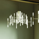TheLight LED 18 Light Chandelier in white, with slim arms radiating from a fluted central cylinder, each carrying glowing vertical LED rods, shown against an olive-toned ceiling.