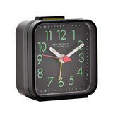 Square plastic alarm clock in black with a top shut-off button, featuring green Arabic numerals, white hands, and red and yellow accent hands on a black dial, shown on a plain white background.