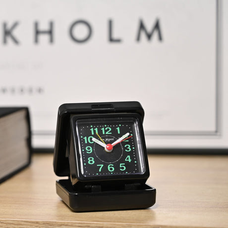 Square black plastic travel alarm clock with folding case open, luminous dial, green Arabic numerals, and white hands, shown on a wooden surface.