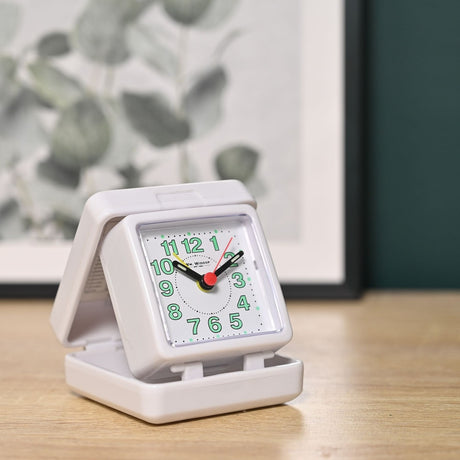 Square folding travel alarm clock in white plastic, open on its hinged base with a white dial, pale green Arabic numerals, black hands and red and yellow centre hands, shown on a wooden surface.
