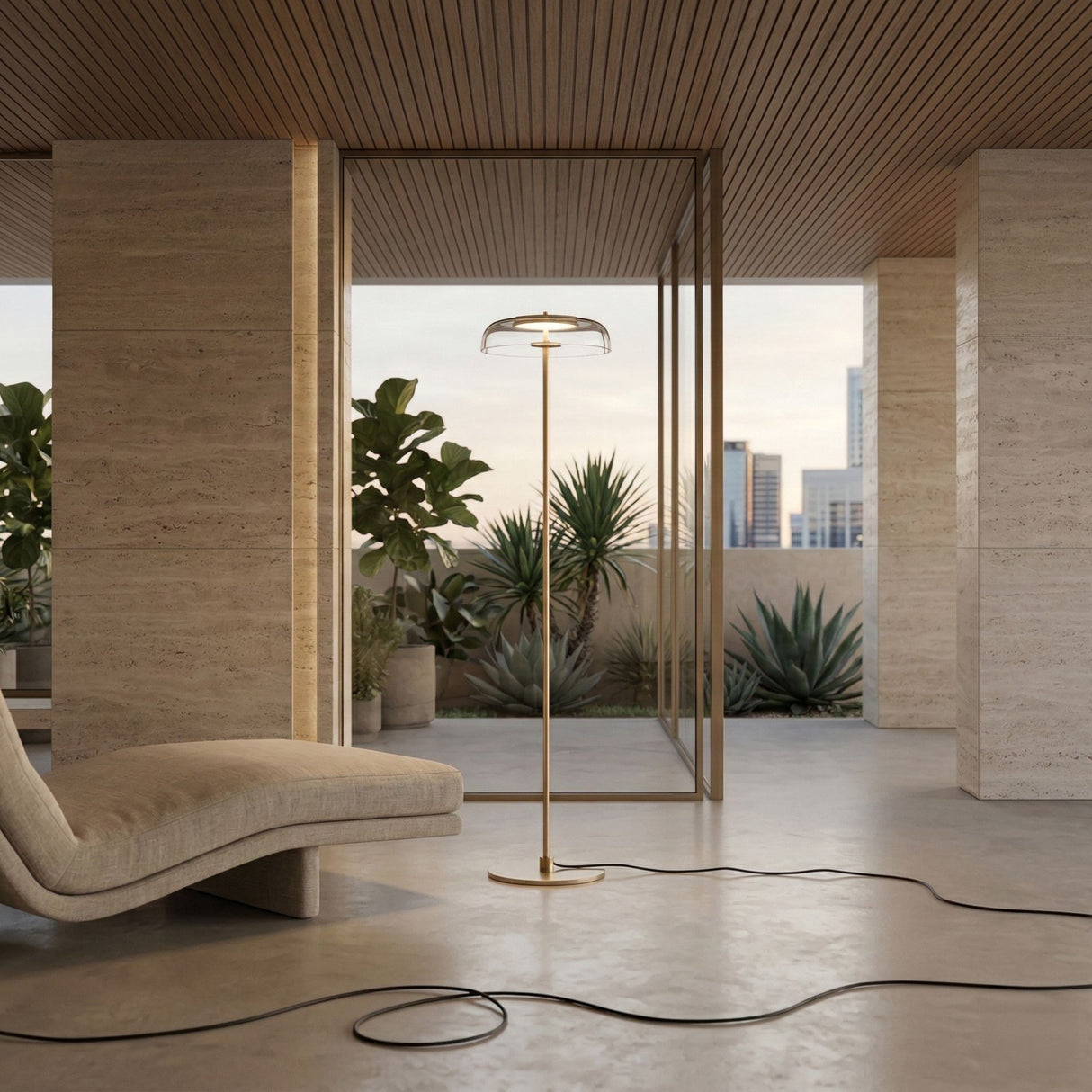 A modern living space features the Blossom LED Floor Lamp in gold, a curved beige lounge chair, large glass doors, and potted plants on a stone-tiled balcony. A coiled black cord rests on the smooth floor.