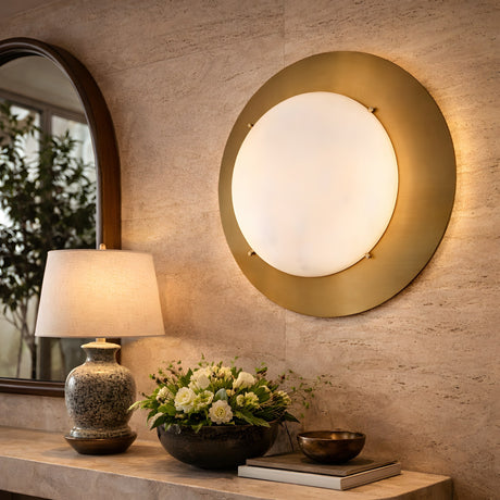 The Lunara 1 Light Round Alabaster Wall Light – Brushed Brass is mounted above a console table styled with a ceramic lamp, flowers, decorative bowl, and a mirror reflecting leafy plants.