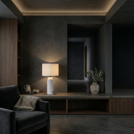 A modern, dimly lit living room showcases a black armchair with a brown throw, the Arvello 1 Light Minimal Marble Table Lamp - White with a beige shade, a ceramic vase of greenery, and dark wood shelves against gray walls and floors.