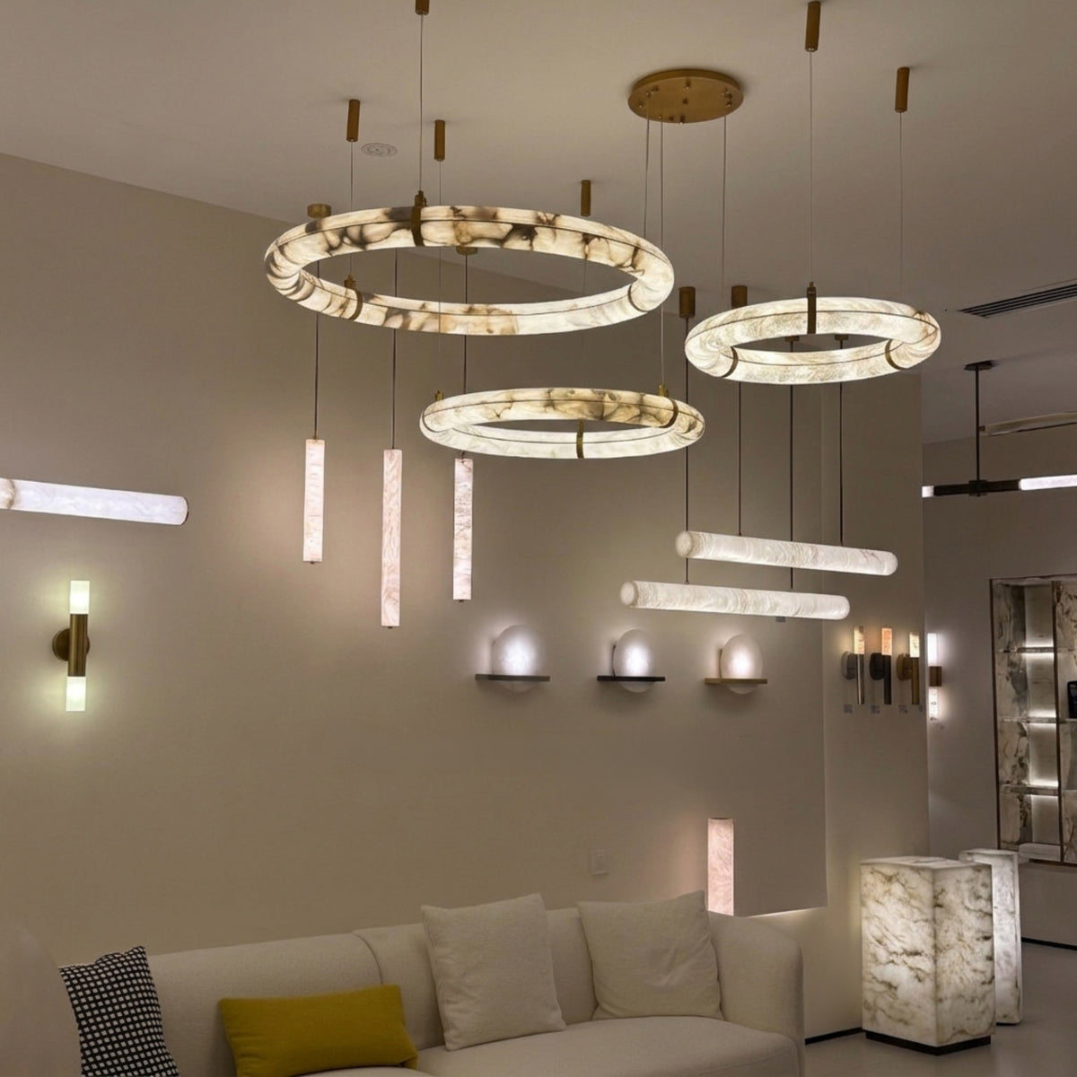 Aureon LED Triple Ring Alabaster Chandelier in brushed brass, three staggered alabaster hoops glowing warm white, shown alongside matching wall and floor lights in a living room setting.