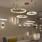 Aureon LED Triple Ring Alabaster Chandelier in brushed brass, three staggered alabaster hoops glowing warm white, shown alongside matching wall and floor lights in a living room setting.