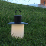 Nautic LED rechargeable outdoor table lamp in anthracite, lantern shape with curved hook handle and flat brim above a glowing milky white cylindrical shade, set in grass at dusk.