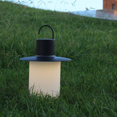 Nautic LED rechargeable outdoor table lamp in anthracite, lantern shape with curved hook handle and flat brim above a glowing milky white cylindrical shade, set in grass at dusk.