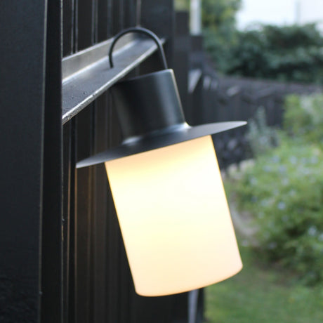 Nautic LED rechargeable outdoor table lamp in anthracite, lantern-style with curved hook handle and wide brim, glowing milky white cylindrical shade, shown hanging outdoors on a dark fence.
