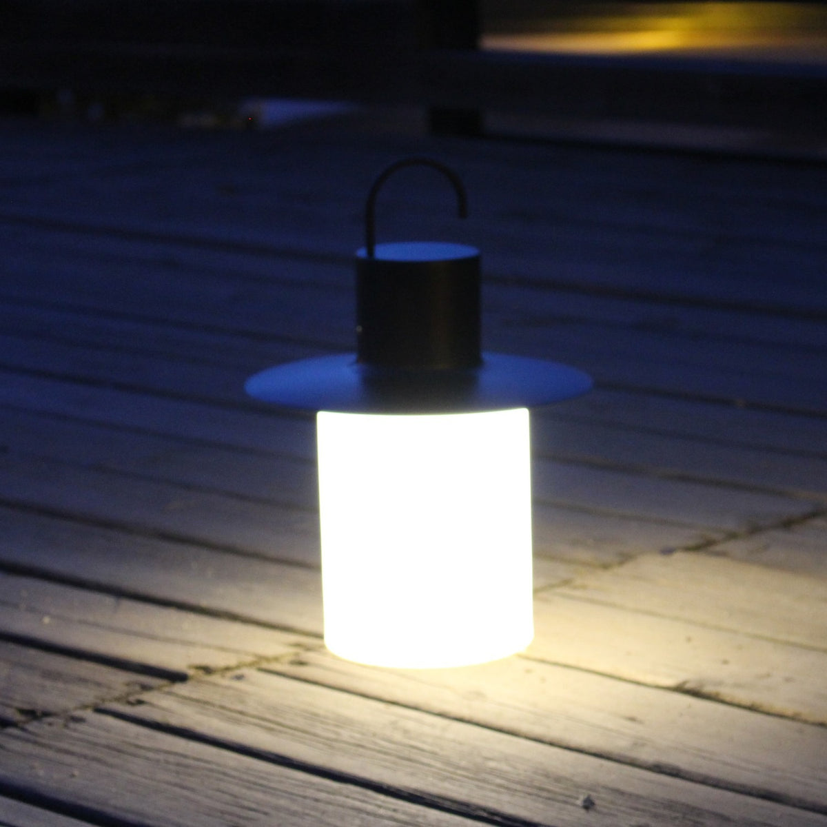 Nautic LED rechargeable outdoor table lamp in anthracite, lantern shape with curved hook handle and wide brim above a glowing white cylindrical shade, shown lit on an outdoor wooden deck at night.