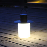 Nautic LED rechargeable outdoor table lamp in anthracite, lantern shape with curved hook handle and wide brim above a glowing white cylindrical shade, shown lit on an outdoor wooden deck at night.