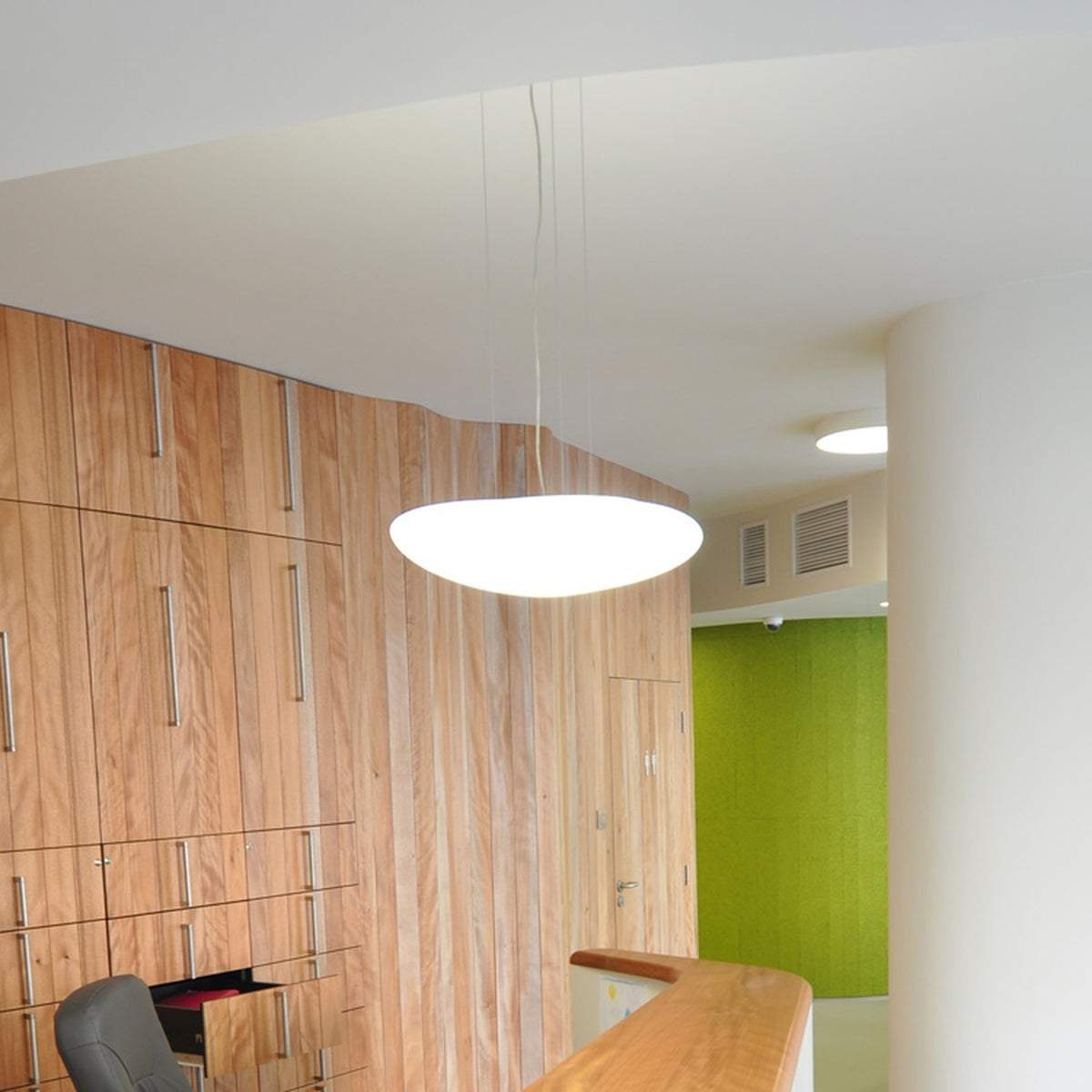 Stone LED Pendant Light in white, a broad flat oval shade suspended on slim wires, shown illuminated above a reception desk in a wood-panelled interior.