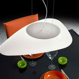 Stone LED Pendant Light in white, a flat pebble-shaped asymmetric shade hung above a glass dining table set with an orange chair and green bowls.
