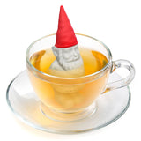 Bearded garden gnome plastic tea infuser in grey and red, steeping in a glass teacup of amber tea on a saucer against a white background.
