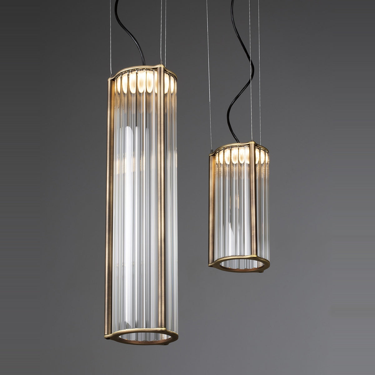 Kalvenne LED glass pendant lights in two sizes, cylindrical ribbed glass columns in satin bronze iron frames, suspended on thin cables against a grey background.
