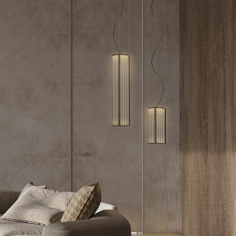 Kalvenne LED Glass Pendant Lights in black, two ribbed glass cylinders in slim black iron frames, shown lit at different heights beside a wood-panelled wall and sofa.
