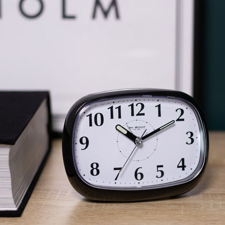 Oval Silent Plastic Alarm Clock in glossy black with white dial, bold black numerals and luminous hands, shown on a wooden surface beside a book.