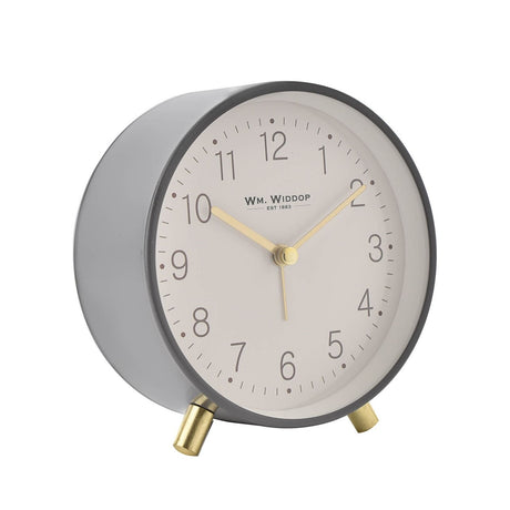 Round alarm clock with a cream dial, Arabic numerals and gold hands, set in a grey metallic case standing on two gold metal feet, shown on a white background.