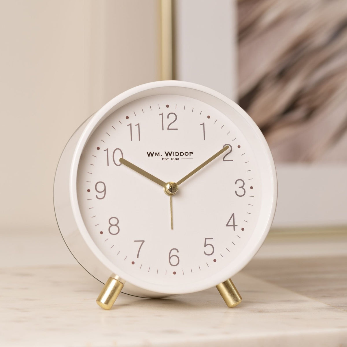 Round white alarm clock with matt metal case, gold feet, and gold hour, minute and second hands, shown on a marble surface against a neutral background.