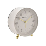 Round white alarm clock with matt metal case, Arabic numeral dial, gold hour, minute and second hands, and slim gold feet, shown on a plain background.