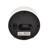 Base view of the round white alarm clock showing the matt black underside with on/off switch, light button, battery compartment, gold metal feet, and product label.