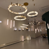 Aureon LED Triple Ring Alabaster Chandelier in brushed brass, shown with matching pendant and wall lights in a hallway setting with warm white ambient lighting.