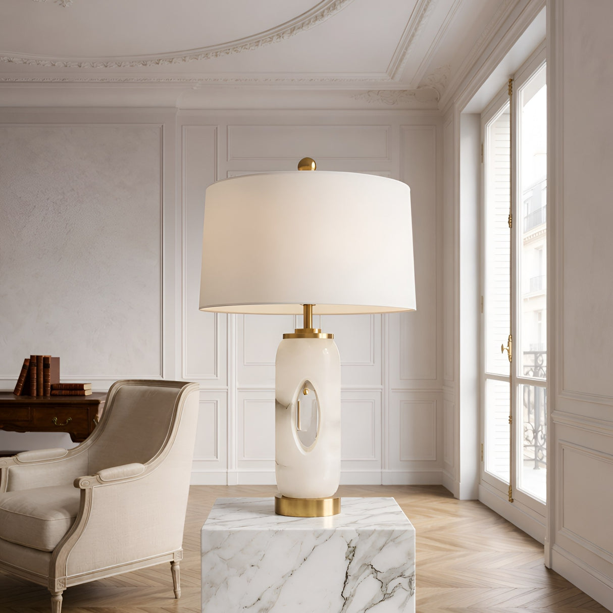 The Elovier 1 Light Carved Oval Alabaster Table Lamp in brass and soft white sits on a marble pedestal in an elegant room with white paneled walls, a large window, and a beige armchair.
