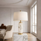 The Elovier 1 Light Carved Oval Alabaster Table Lamp in brass and soft white sits on a marble pedestal in an elegant room with white paneled walls, a large window, and a beige armchair.